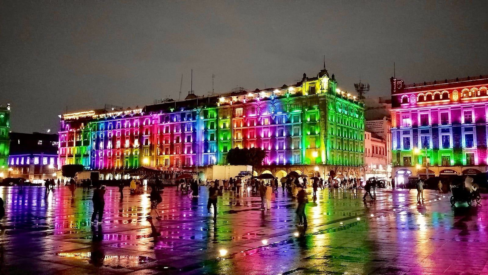 Night view of Mexico City’s Zócalo with historic buildings lit up in colorful lights and reflections on the wet plaza.