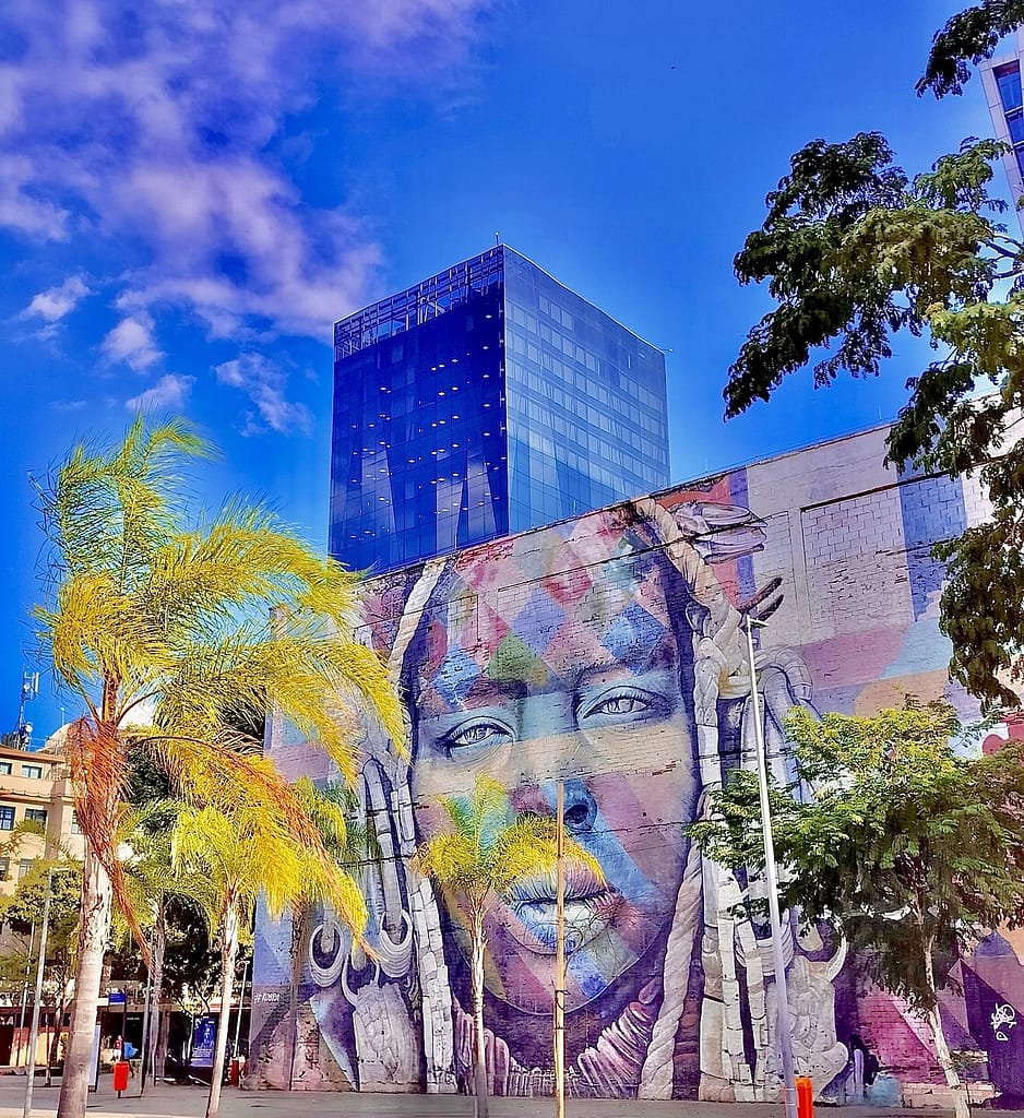Discover Olympic Boulevard in Rio de Janeiro: Street Art, Waterfront Strolls & Cultural Highlights 18 Panoramic view of Eduardo Kobra’s “Etnias” mural on Olympic Boulevard in Rio de Janeiro, with a modern glass building and palm trees under a clear blue sky.